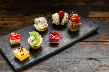Selection of different dessert pastry sweet mini cakes and tartlets with cream and fruit on slate and wooden background