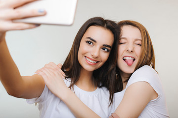 Beautiful young caucasian brunette doing selfie smiling while her female friend is hugging her and showing teeth with closed eyes isolated on white background.