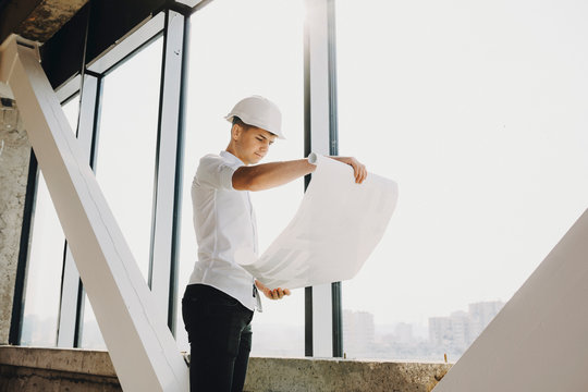 Confident adult arhitect looking at the plan of the building in construction near a big window.