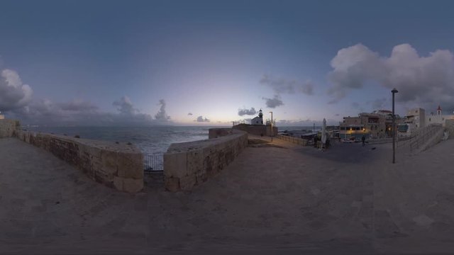 360 VR video. Acre view in late evening. Old coastal city with houses, ancient walls and active lighthouse. Man tourist walking in the street