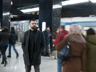 Handsome bearded man dressed in wool coat stands still in metro station within moving crowds of...
