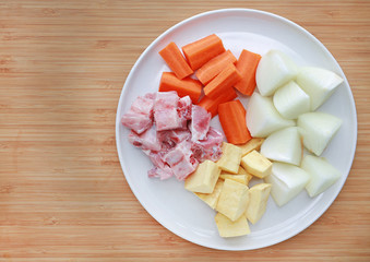 Sliced carrot, onion, pork bone and potato on white plate against wooden background Ingredients for boiling soup.