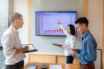 Beautiful businesswoman gives report or presentation to her business colleagues in the conference room. She show graphic charts and company's growth on the wall TV.