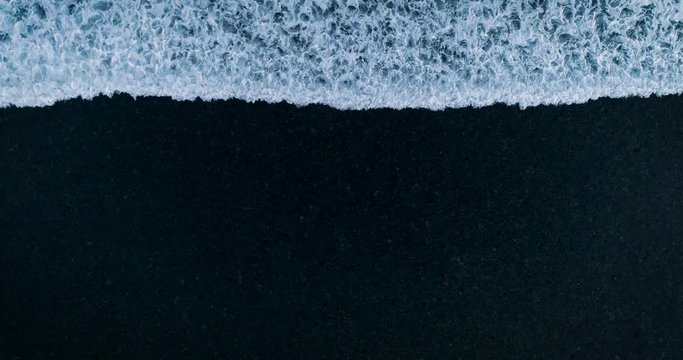 Aerial bird's eye view white foam wave gradually dissolves and disappears on the black sand. Сoncept looped background of disappearance, dissolution 4K