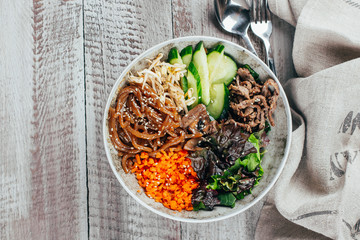 Asian buddha bowl on wooden table background. Grilled beef, carrot, cucumber, sprouted mung bean, topinambour noodles, mushrooms, sesame seeds, asian sause. Top view, copy space.