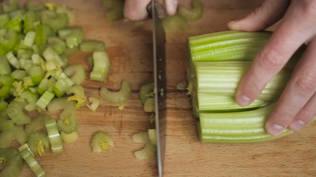 Knife chopping celery in restaurant kitchen on wooden cutting surface. Chef cutting celery to cook in the kitchen. Hands Slicing celery with Knife on chopping wooden board background. Slow motion.