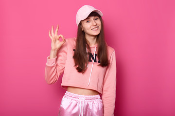 Obraz na płótnie Canvas Close up photo of pretty attractive lady wearing rosy sport pullover, sweatpants, cap, shows okay sign, model poses isolated over bright pink background. Teenager, style and fashion concept.