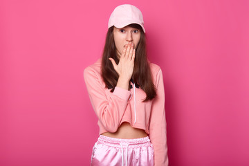 Portrait of surprised European brunette teenager covers her opened mouth with hand, dressed in stylish rose sweatpants, hoodie and cap, stands against pink studio wall. People and emotions concept.