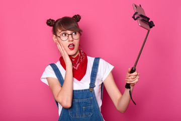 Close up portrait of attractive female making selfie, using selfie stick, poses, takes photo for her social network, makes surprised look, stands with open mouth, keeps hand on cheek. Teen concept.