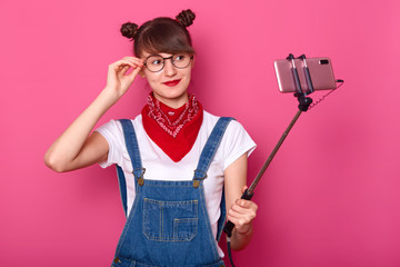 Attractive woman stands alone over pink background, making selfie while trying on new stylish glasses, uses selfie stick, looks smiling at camera, keeps hand on spectacles. Girl buys trendy eyewear.