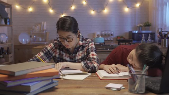 female college student fall asleep at desk in dining room with kitchen in background. lazy girl sleep on table while another smart woman in glasses hard working writing doing homework at night home.