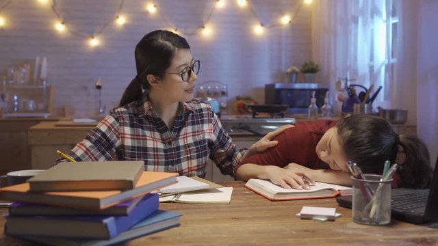 smart young college girl student wake her roommate up to keep study in kitchen table. young woman get up and stretch body sitting at desk at night home. two friends doing homework together indoors.