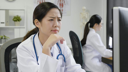 Young asian woman doctor sitting at working desk looking computer screen with serious face thinking...
