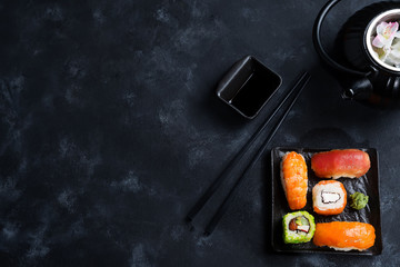 Asian food background with black iron teapot and sushi set on slate plate on black stone table, flat lay