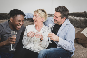 An interracial gay couple, with extended family, sitting on a sofa at home and celebrating with a glass of champagne. Black, Caucasian and Mixed race people. 
