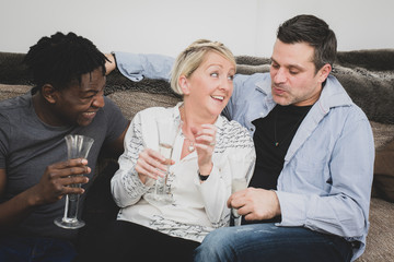 An interracial gay couple, with extended family, sitting on a sofa at home and celebrating with a glass of champagne. Black, Caucasian and Mixed race people. 