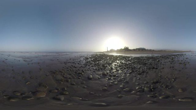 GRAN CANARIA, CANARY ISLANDS - AUGUST 02, 2016: 360 VR Video. People on vacation walking along the ocean coast with rocks. View to the resort with Maspalomas Lighthouse against setting sun