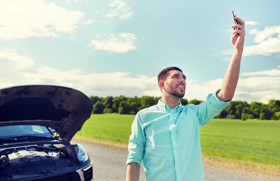 road trip, transport, travel, technology and people concept - young man with smartphone catching signal and open hood of broken car at countryside