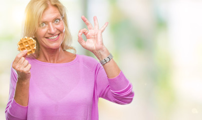 Middle age blonde woman eating sweet waffle over isolated background doing ok sign with fingers, excellent symbol