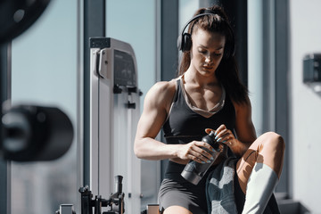 Young Caucasian strong muscular female bodybuilder sitting in gym with headphones on ears and water...