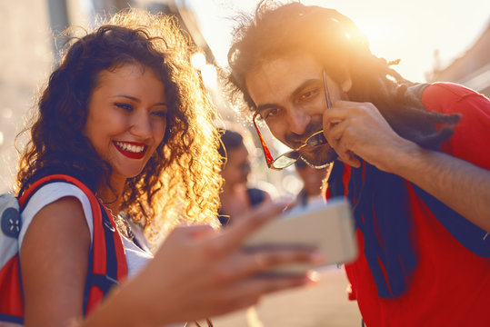 Cute smiling young woman with curly hair and backpack on backs showing to her mixed race friend something on smart phone. Summer vacations concept. - Powered by Adobe