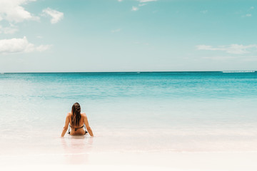 Woman with long brown hair and in swimsuit sitting in shoal and enjoying sunny weather and ocean. Backs turned.