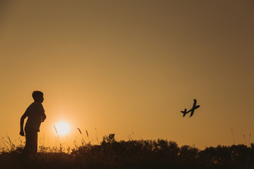 Black small silhouette of young happy kid playing toy plane outdoors at sunset time. Horizontal color photography.