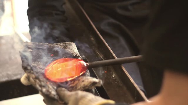 Male close up in glass blowing workshop, rolling molten hot orange glowing glass into formed shape on heat proof pad.