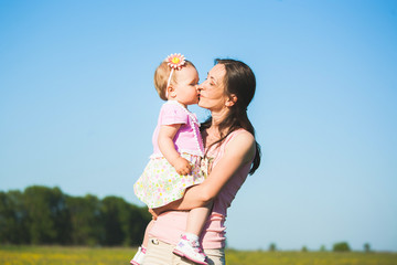 Young mother kisses her little toddler daughter holding her in hands while standing at sunny spring meadow outside at countryside. Horizontal color photography.