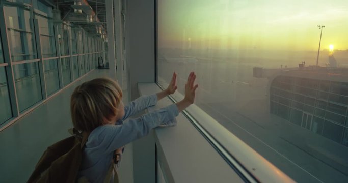 Cute small caucasian boy with a backpack standing at the big window in the airport and looking outside at the planes and beautiful sunrise. Rear.