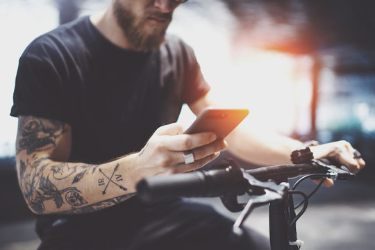 Bearded tattooed man in sunglasses using mobile phone for send text message after riding by electric scooter in the city