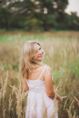Fototapeta na wymiar Portrait of smiling happy blond young beautiful girl standing outside in grassy meadow and looking back. Horizontal color photography.