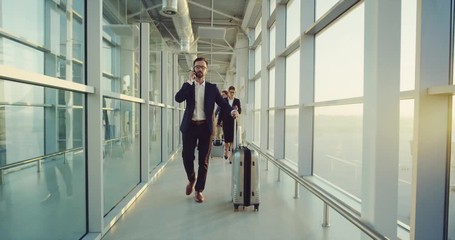 Caucasian handsome businessman in the costume going by the corridor of the airport with a suitcase and talking on the phone. - Powered by Adobe