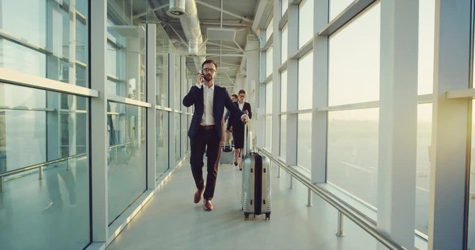 Caucasian handsome businessman in the costume going by the corridor of the airport with a suitcase and talking on the phone.