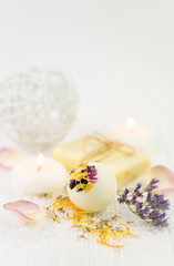 Beautiful set with selective focus on white creamy moisturizing small bath ball bomb with dry flower blossom petals, spa candles burning and dry flower pieces scattered on white clean soft towel.