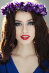 Naklejka premium Fashion Portrait of a beautiful brunette woman with amazing make up and coronet of purple flowers in her head, smiling at camera, outside background. Vertical view.