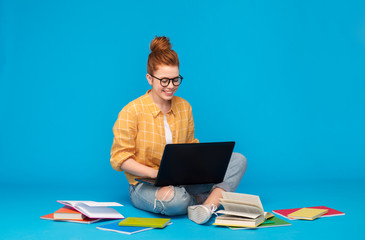 education, high school, technology and people concept - red haired teenage student girl in checkered shirt and torn jeans with books using laptop computer over bright blue background - obrazy, fototapety, plakaty