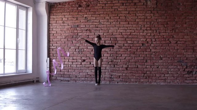 Caucasian little girl in black body perform gymnastics exercise with a pink ribbon in studio with windows and brick wall background at gymnastics school. Workout, slow motion