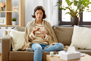 people, health and treatment concept - sad sick young woman in blanket taking medicine with water at home