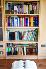 Open book with glasses on blurry background of bookshelves with colorful books. Library, reading, learning concept.