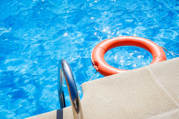 Closeup view of the swimming pool edge with staircase and a red ring life buoy floating on the blue water during a sunny summer day