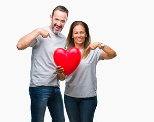 Fototapeta na wymiar Middle age hispanic casual couple in love holding red heart over isolated background with surprise face pointing finger to himself