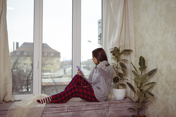 girl teenager sits on the window with the phone in hand and listens to something in the headphones. Loneliness. A young woman looks out the window, her face is not visible.