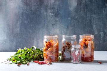 Korean traditional fermented appetizer kimchi cabbage and radish salad, hot spicy anchovies fish snack served in glass jars with Vietnamese oregano greens and chili peppers over grey blue table. - obrazy, fototapety, plakaty