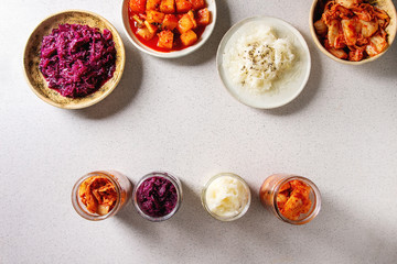 Variety of fermented food korean traditional kimchi cabbage and radish salad, white and red sauerkraut in glass jars and ceramic plates over grey spotted background. Flat lay, space