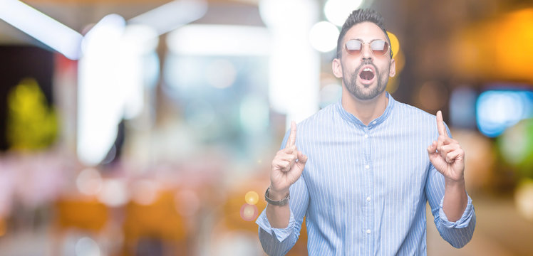Young handsome man wearing sunglasses over isolated background amazed and surprised looking up and pointing with fingers and raised arms.