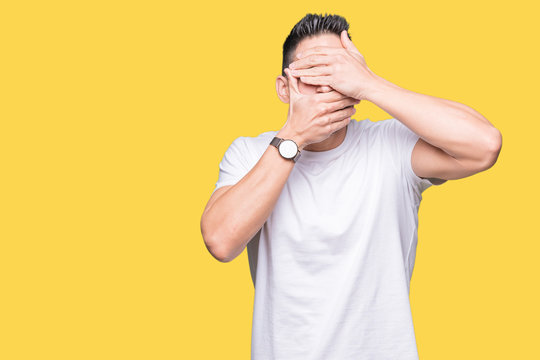 Handsome man wearing white t-shirt over yellow isolated background Covering eyes and mouth with hands, surprised and shocked. Hiding emotion