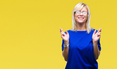 Young beautiful blonde woman wearing glasses over isolated background smiling crossing fingers with hope and eyes closed. Luck and superstitious concept.