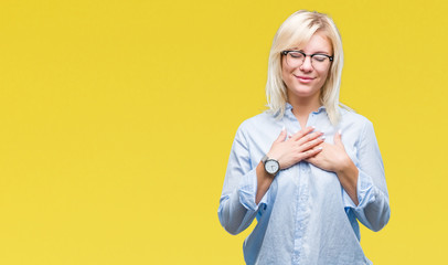 Young beautiful blonde business woman wearing glasses over isolated background smiling with hands on chest with closed eyes and grateful gesture on face. Health concept.
