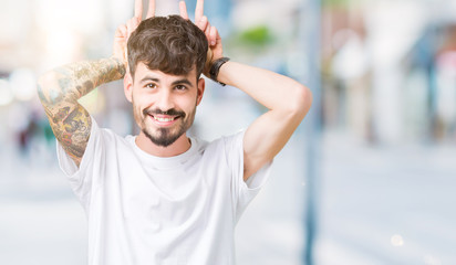 Fototapeta premium Young handsome man wearing white t-shirt over isolated background Posing funny and crazy with fingers on head as bunny ears, smiling cheerful